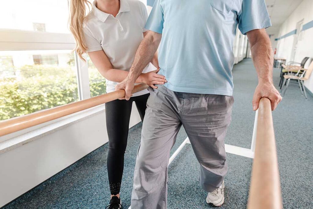 Physical therapist guiding an adult through a balance exercise beside parallel bars.