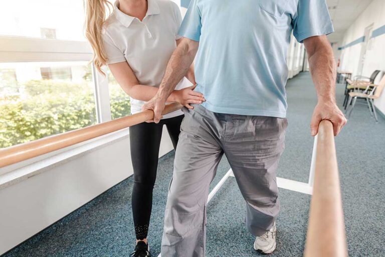 Physical therapist guiding an adult through a balance exercise beside parallel bars.